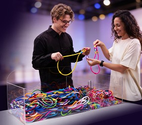 Un homme et une femme créant leurs propres lanyards publicitaires devant le présentoir transparent MIX'n'MATCH Experience Box sur un stand d'exposition.